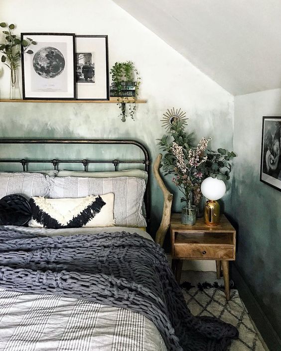 a cozy attic bedroom with green watercolor ombre walls, a metal bed with printed bedding, a ledge with potted plants and art