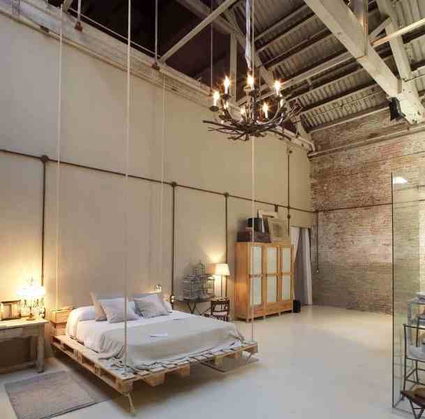 a creative neutral bedroom with industrial touches - a roof, brick walls, a suspended pallet bed, a wooden wardrobe and an antler chandelier, some lamps