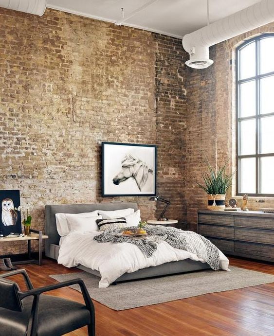 a light-flooded industrial bedroom with brick walls, a reclaimed wood dresser, a bench, a grey upholstered bed with neutral bedding, exposed piping