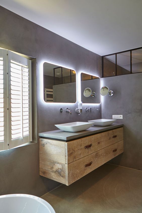 a minimalist industrial bathroom done of concrete and rough wood, with a floating vanity, lit up mirrors, sinks and shutters