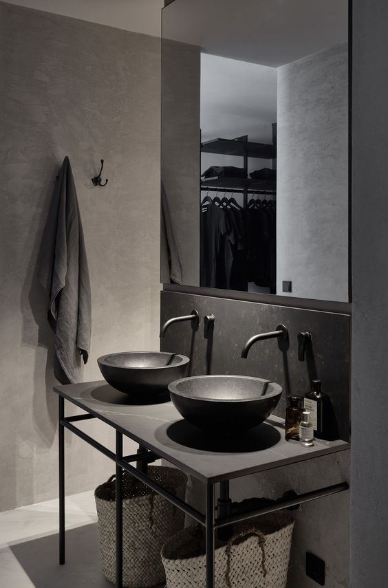 a minimalist industrial bathroom with concrete and stone walls and a floor, a vanity with stone bowls, a mirror and baskets