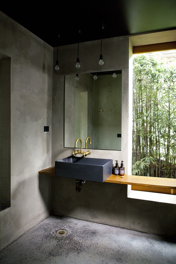 a minimalist industrial bathroom with concrete walls and a floor, a wooden vanity and a stone sink, exposed pipes and bulbs