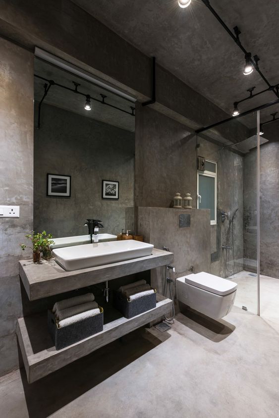 a minimalist industrial bathroom with concrete walls and a floor, exposed pipes and lamps, a large mirror and a floating vanity of concrete