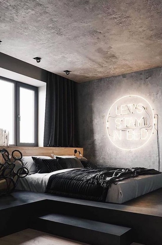 a modern industrial bedroom with a concrete ceiling and walls, a neon light, a platformbed and dark textiles