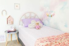 a pastel kid’s rom with a pastel watercolor accent wall, a pink bed with pastel bedding, a pink rug, a side table and some decor