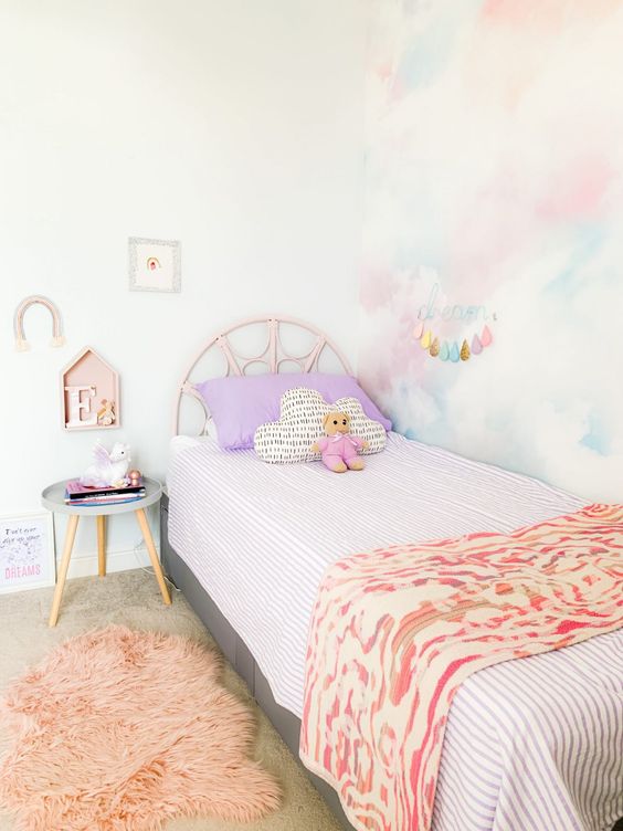 a pastel kid's rom with a pastel watercolor accent wall, a pink bed with pastel bedding, a pink rug, a side table and some decor