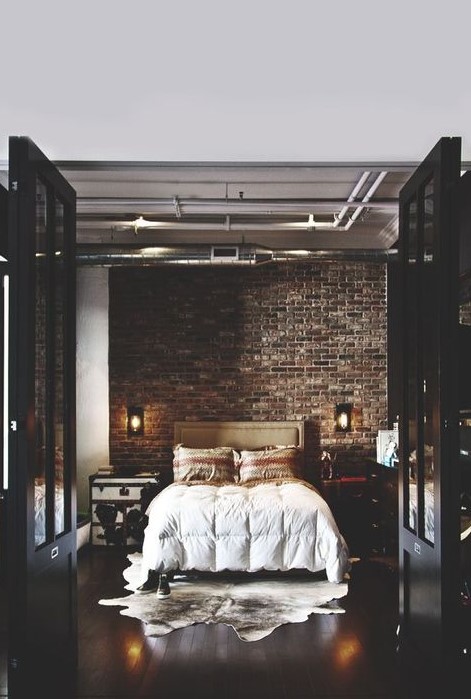 a refined industrial bedroom with brick walls, an upholstered bed, a chest and a dresser and exposed pipes on the ceiling