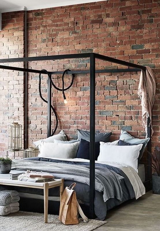 a simple industrial bedroom with red brick walls, a black metal frame bed, wooden and wicker furniture and cozy textiles