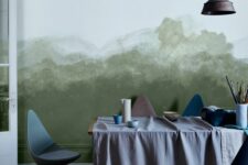 a stylish art or dining room with a green watercolor wall, a table with paints and brushes, blue chairs and a metal pendant lamp