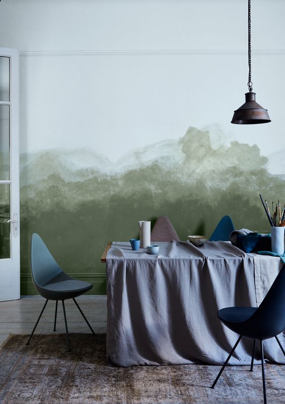 a stylish art or dining room with a green watercolor wall, a table with paints and brushes, blue chairs and a metal pendant lamp