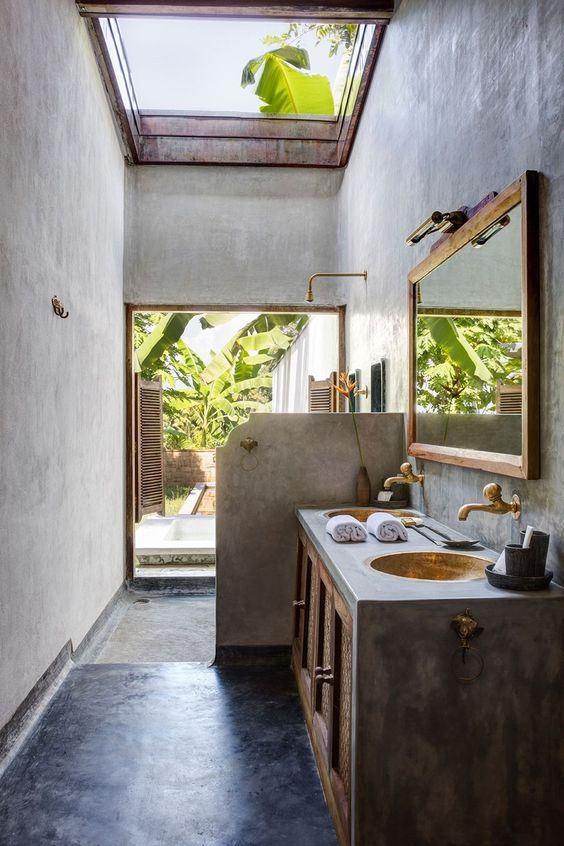 a tropical industrial bathroom with concrete walls and a floor, a concrete vanity and a half wall, metal sinks and gold faucets