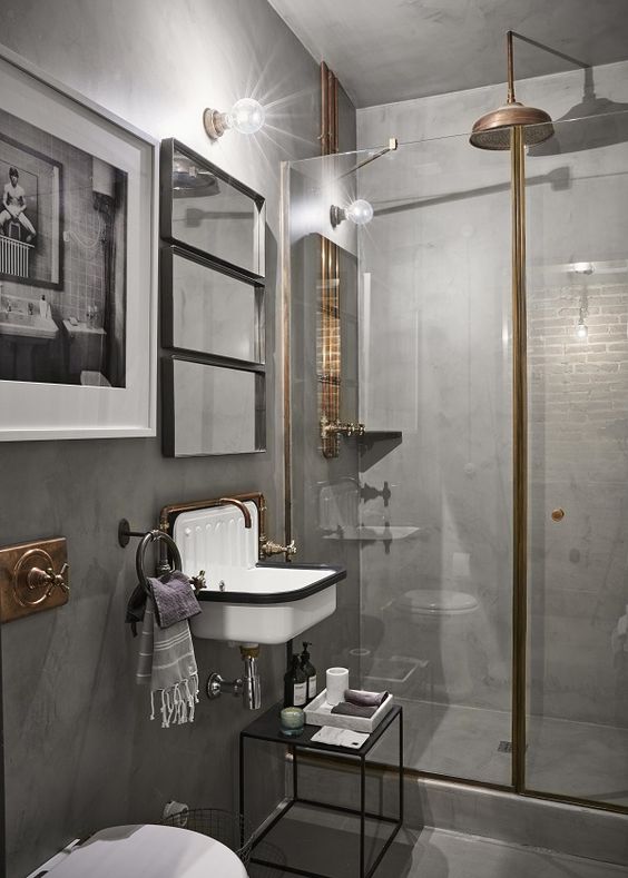 a vintage industrial bathroom clad with concrete, with a vintage sink, artworks and a shower enclosed in glass and exposed pipes
