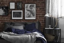 a welcoming industrial bedroom with a red brick accent wall, a bed with grey bedding, a crock bench, a black metal nightstand on casters and a gallery wall