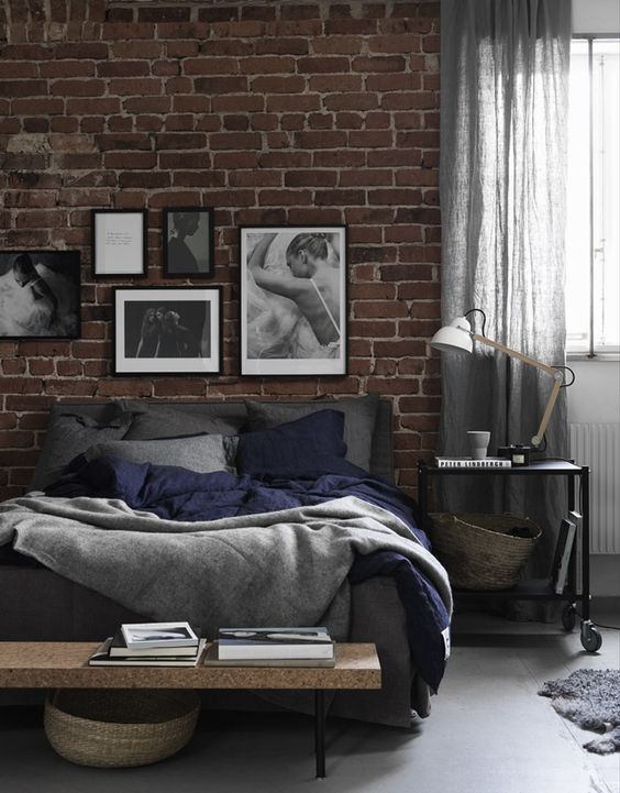 a welcoming industrial bedroom with a red brick accent wall, a bed with grey bedding, a crock bench, a black metal nightstand on casters and a gallery wall