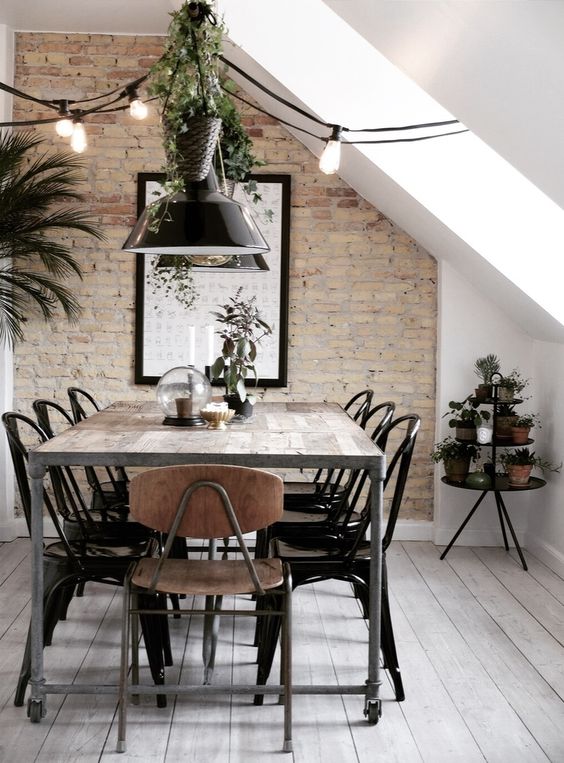 an industrial Scandinavian dining room with a brick accent wall, a metal and wood table, metal chairs, black pendant lamps and greenery