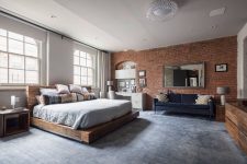 an industrial bedroom with a red brick wall, a wooden slab bed with neutral bedding, a white desk and a grey chair plus a crystal chandelier