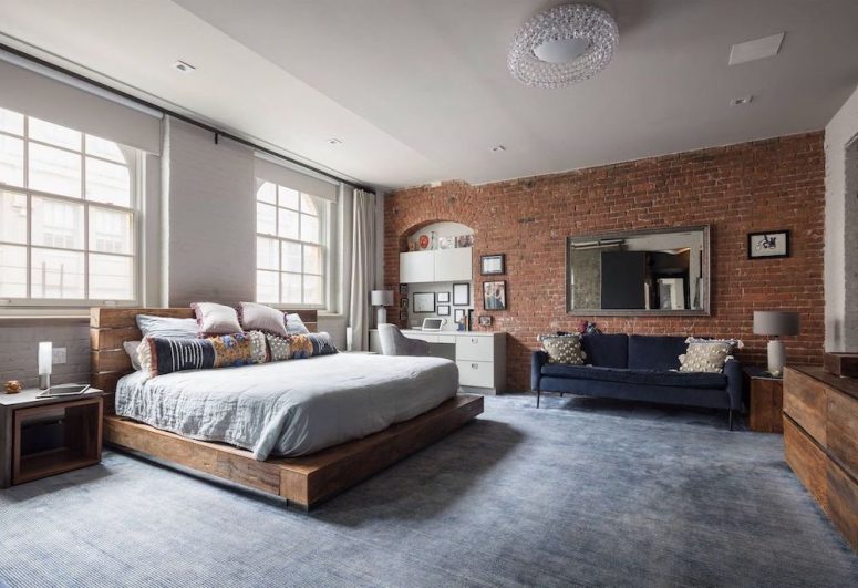 an industrial bedroom with a red brick wall, a wooden slab bed with neutral bedding, a white desk and a grey chair plus a crystal chandelier