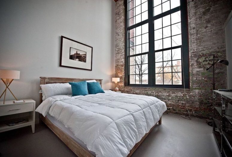 an industrial bedroom with a shabby chic brick wall, a wooden bed with neutral bedding, a white nightstand, double-height windows for more light