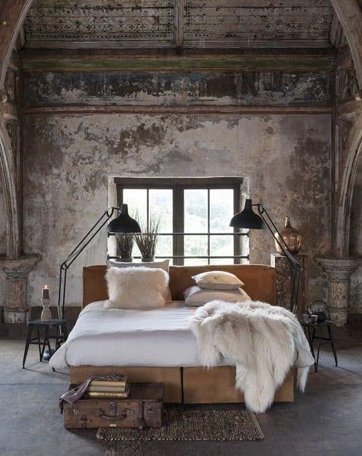 an industrial bedroom with shabby chic walls, a wooden ceiling, a concrete floor and upholstered bed, metal floor lamps and a vintage suitcase