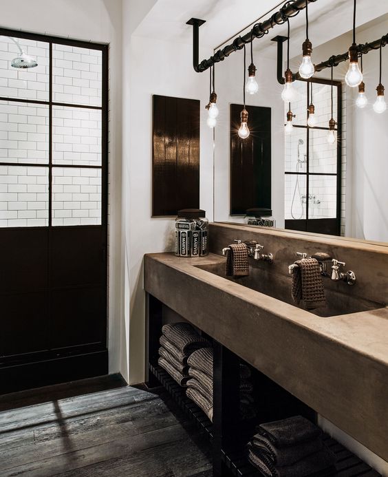 an industrial chic bathroom with a glazed shower space, a concrete vanity with a double sink, a rough wooden floor, exposed pipes and bulbs