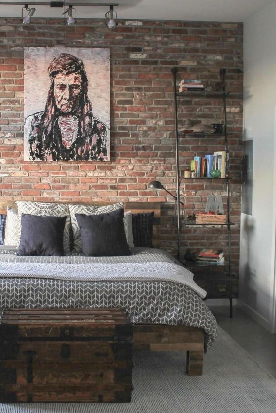an industrial sleeping space with a red brick wall, a wooden bed and a wooden chest for storage, a black metal ladder and some books