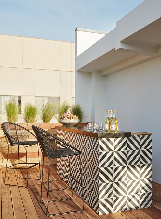 an outdoor bar with a tiled black and white bar countertop and black stools and potted greenery and lots of drinks