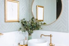 a beautiful bathroom with pale blue chevron tiles, a white vanity, gold handles, a round mirror in a gold frame, gold fixtures and an artwork in a gold frame