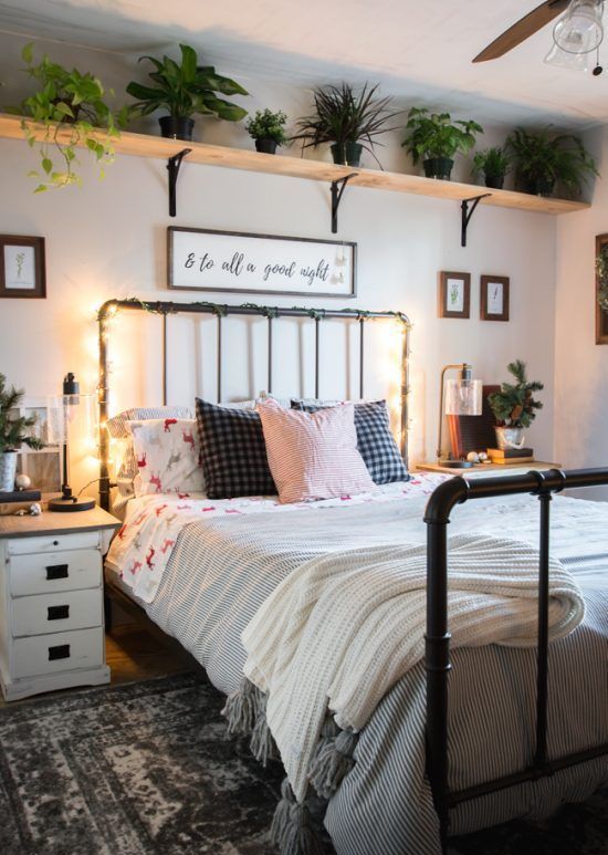 a bedroom with a metal pipe bed, neutral bedding, a wall mounted shelf, white nightstands, table lamps and potted plants