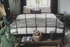 a boho bedroom with a black metal bed with neutral bedding, a printed rug, a wooden bench, potted plants, tassels and hangings