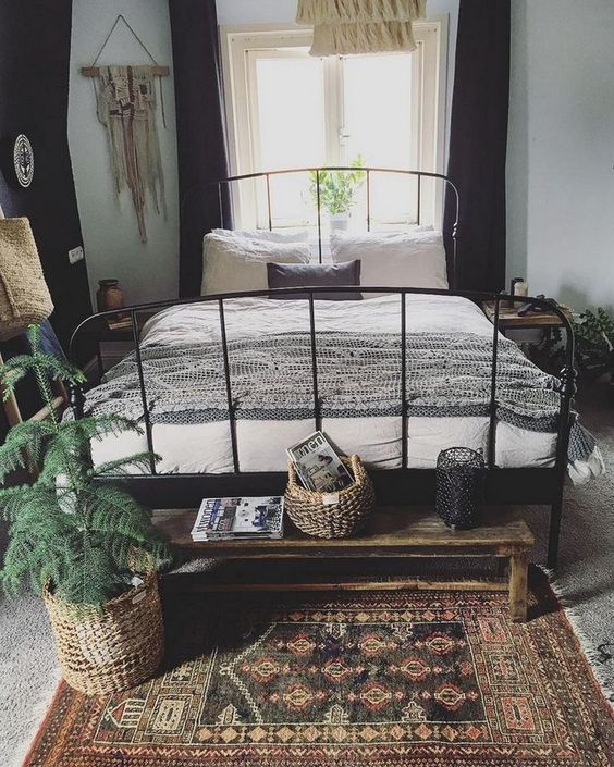 a boho bedroom with a black metal bed with neutral bedding, a printed rug, a wooden bench, potted plants, tassels and hangings