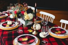 a bold Thanksgiving tablescape with a plaid blanket and plates, tall candles, fall leaves and pumpkins plus florals