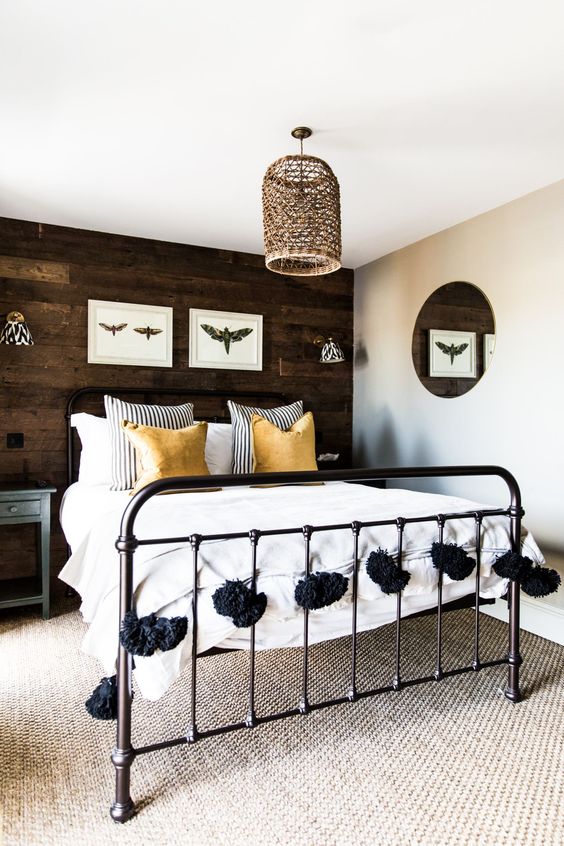 a farmhouse bedroom with a dark stained accent wall, a black metal bed with neutral bedding, wall sconces and a woven lamp