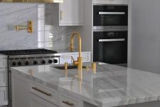 a modern white kitchen with white stone countertops and a backsplash, gold fixtures and a hood lined with gold, gold pendant lamps