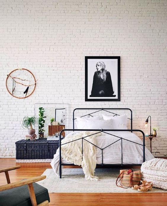 a neutral industrial bedroom with a white brick wall, a black metal bed with neutral bedding, a woven chest and potted plants