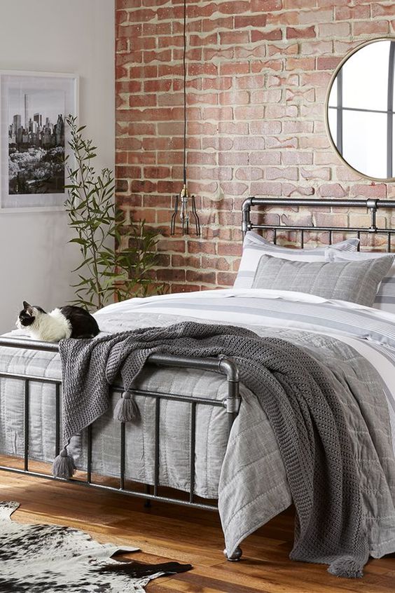 a pretty industrial bedroom with a red brick accent wall, a metal piping bed with grey bedding, pendant lamps and potted plants