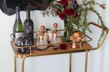 a refined Halloween bar cart with a burgundy and greenery centerpiece, brass and copper skulls, a bat and black paper blooms over it