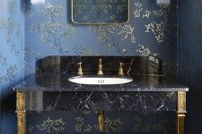 a refined bathroom with navy and gold floral wallpaper, a black marble sink on gold legs and gold fixtures, a mirror in a gold mirror and gold sconces