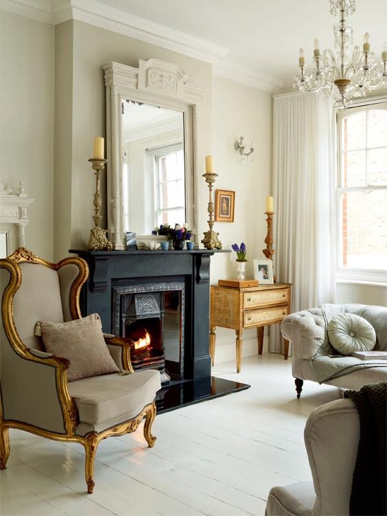a sophisticated living room in neutrals, with a black fireplace, a crystal chandelier, a large mirror, grey furniture, a chair in a gilded frame and gilded candleholders