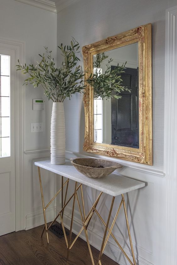 a stylish entryway with a large mirror in a statement gilded frame, a console table with gilded legs, a tall vase and a bread bowl
