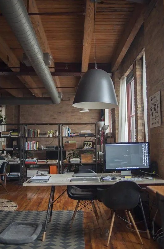 a stylish industrial home office with red brick walls, a comfy desk and a black chair, a metal shelving unit, exposed pipes and metal pendant lamps