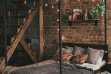 an industrial bedroom with a red brick accent wall, a dark-stained and metal bed with grey bedding, lights and a shelf