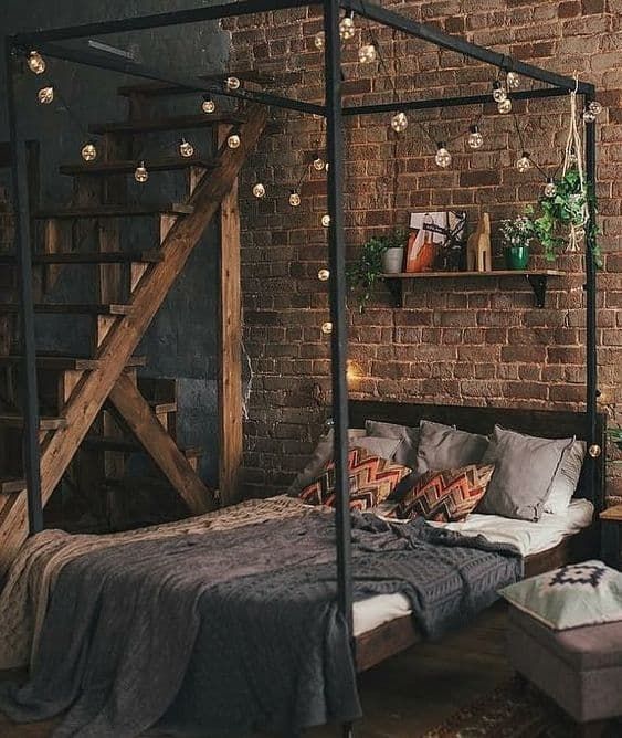 an industrial bedroom with a red brick accent wall, a dark-stained and metal bed with grey bedding, lights and a shelf