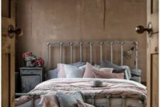 an industrial bedroom with brown walls, a metal bed with grey and pink bedding, a whitewashed nightstand and a metal basket