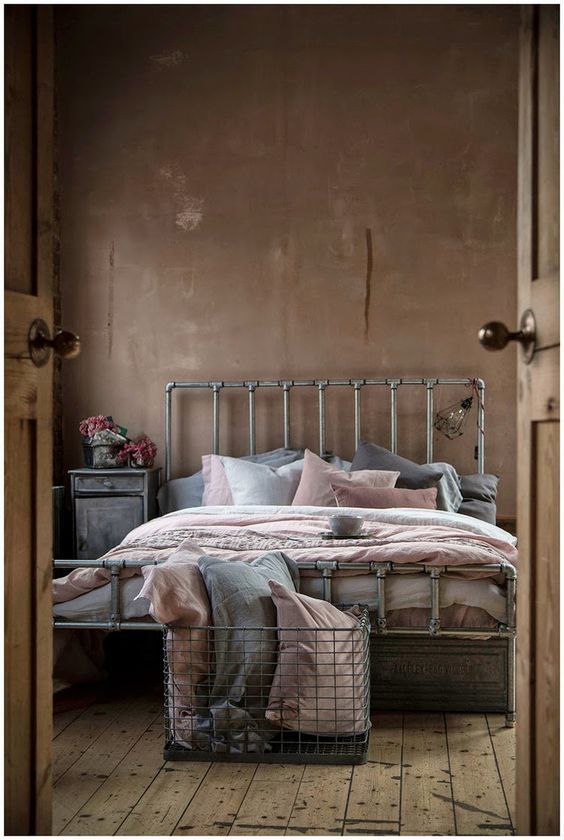 an industrial bedroom with brown walls, a metal bed with grey and pink bedding, a whitewashed nightstand and a metal basket
