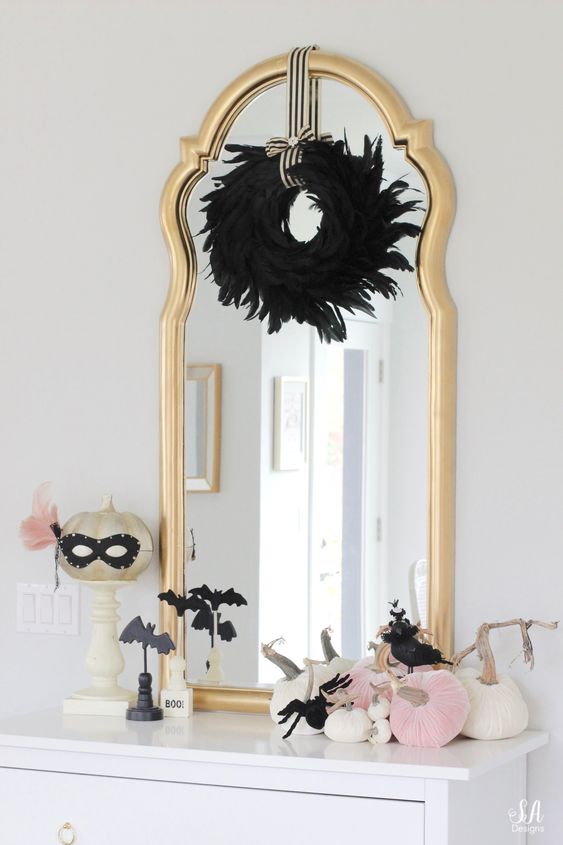 glam Halloween styling with a black feather wreath, a gold pumpkin in a mask, pink and white pumpkins and black bats