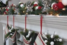 a Christmas mantel decorated with evergreens, white, silver and red ornaments, deer heads, lights, grey embroidered stockings with evergreens