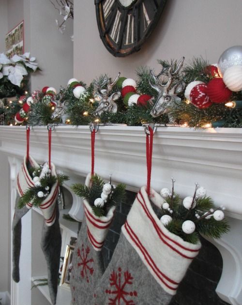 a Christmas mantel decorated with evergreens, white, silver and red ornaments, deer heads, lights, grey embroidered stockings with evergreens