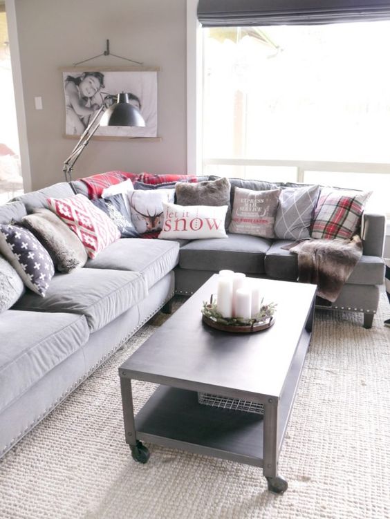 a Christmassy living room with a grey sectional, lots of grey and red printed pillows, a grey table with pillar candles and a lamp