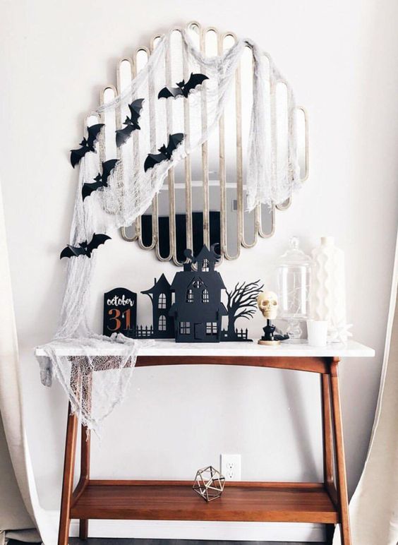 a Halloween console table with cheesecloth, black bats, black houses and a white skull is a bold and cool idea