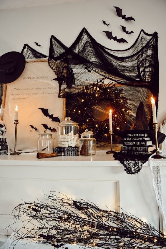 a Halloween mantel styled with black bats, a black wreath with lights, stacks of books, black branches and a black witch hat
